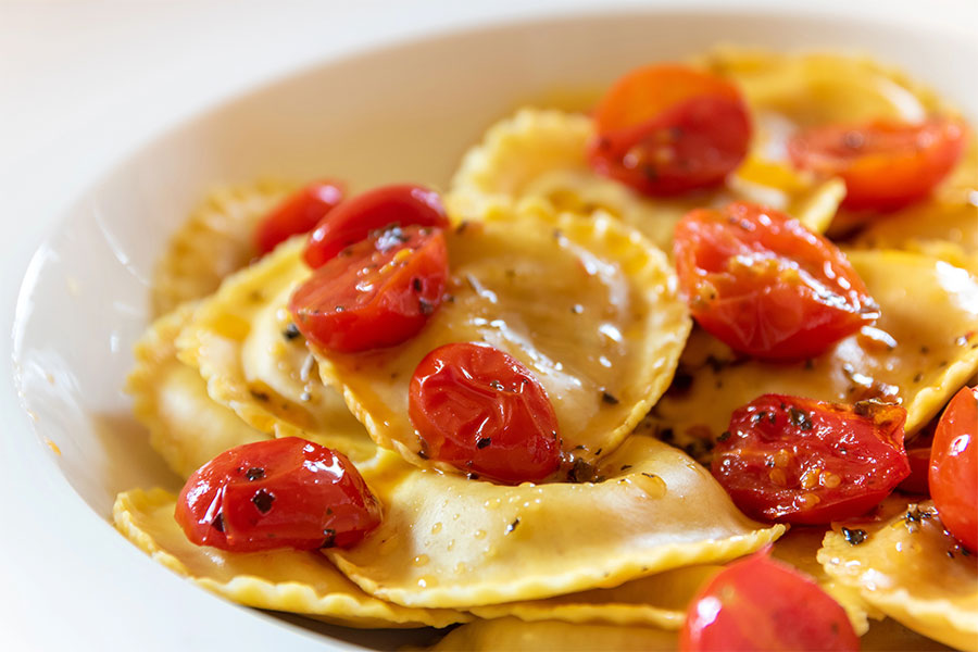 Primo piano di ravioli conditi con pomodorini saltati e un filo d’olio, serviti in un piatto bianco.
